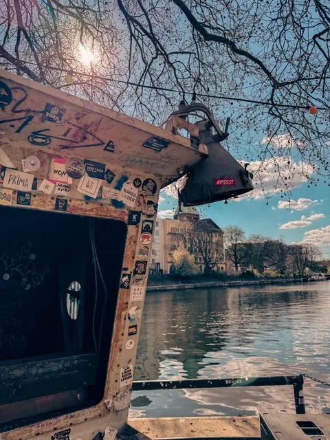 Verwittertes Bootshaus mit Stickern und Graffiti am Spreeufer nahe der Jannowitzbrücke in Berlin, mit Sonnenlicht, kahlen Ästen und Wasser im Hintergrund.