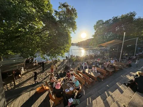 Gut besuchter Uferbereich an der Spree nahe der Jannowitzbrücke in Berlin, mit Sitzplätzen, Bäumen und Sonnenuntergang über dem Wasser.